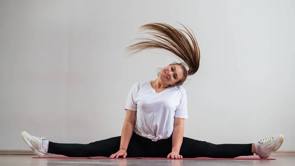 Une jeune femme grasse flexible s'assoit dans une ficelle transversale et agite ses cheveux sur un fond blanc — Photo