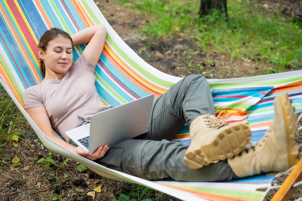 Mulher caucasiana trabalhando em laptop enquanto sentada em uma rede na floresta. Menina usa um computador sem fio em uma caminhada. — Fotografia de Stock