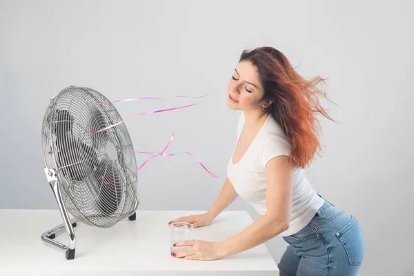 Una mujer caucásica pelirroja se enfría junto al ventilador eléctrico y bebe una bebida fría. Climatización en el apartamento — Foto de Stock