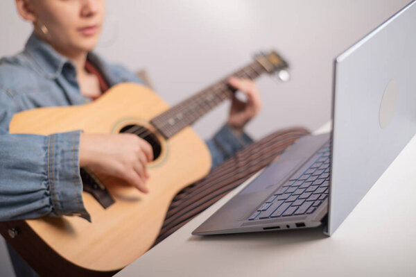 Young caucasian woman with short blonde hair playing guitar and watching training video on laptop.