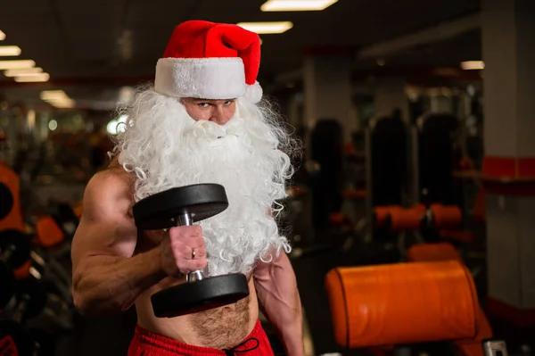 Santa Claus en el gimnasio. Hombre musculoso con un torso desnudo haciendo ejercicios con pesas. —  Fotos de Stock