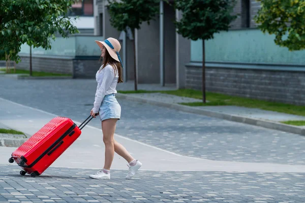 Gelukkig blanke jonge vrouw met een hoed en korte broek houdt een grote rode koffer vast loopt op een straat in de stad — Stockfoto