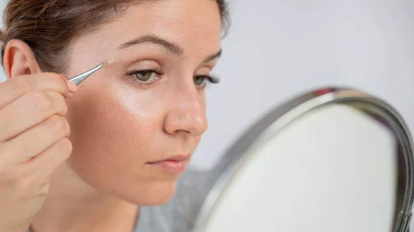 Mujer caucásica se mira en el espejo y hace la corrección de cejas a sí misma con pinzas — Foto de Stock