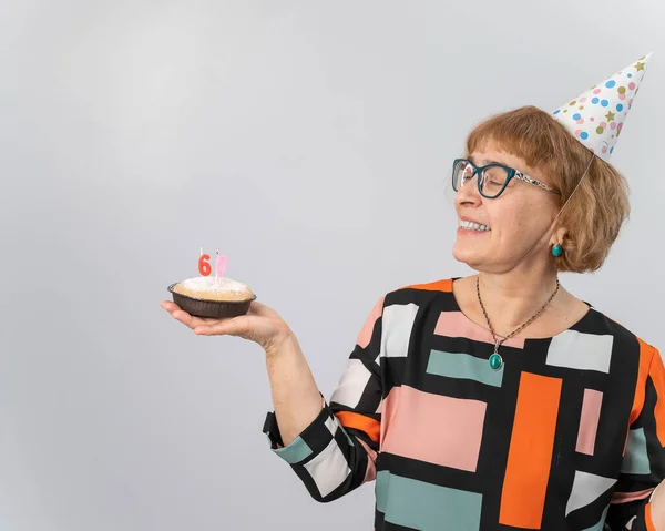 Портрет усміхненої літньої жінки в святковій шапці, що тримає торт зі свічками у вигляді номера 60. ювілею — стокове фото