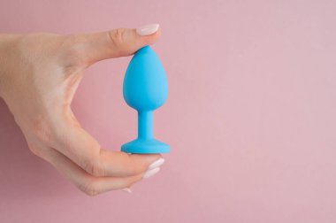 A woman is holding a blue butt plug on a pink background. Adult toy for alternative sex