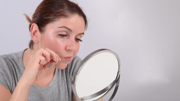 Blanke vrouw kijkt in de spiegel en verwijdert de snor zelf met behulp van een pincet. Hirsutisme. — Stockvideo