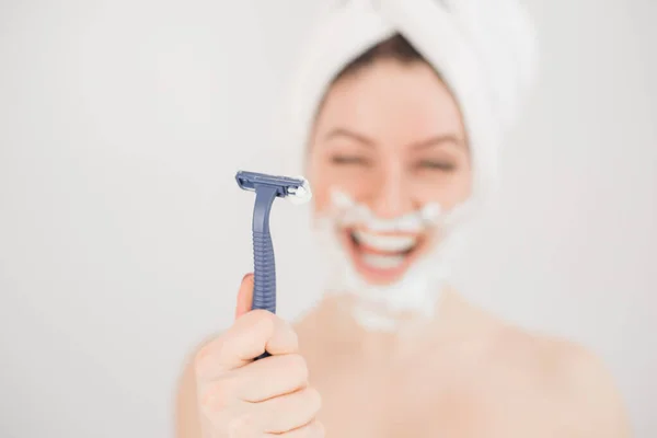 Χαρούμενη καυκάσια γυναίκα με μια πετσέτα στο κεφάλι και αφρό ξυρίσματος στο πρόσωπό της κρατά ένα ξυράφι σε λευκό φόντο — Φωτογραφία Αρχείου