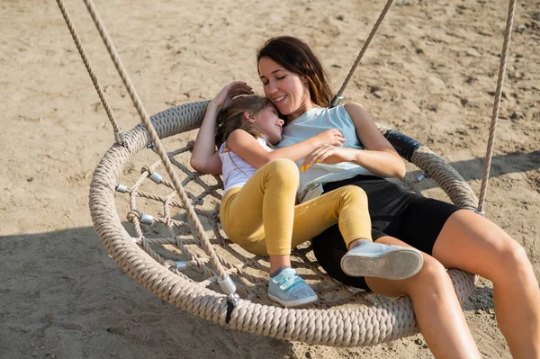 Mutter und Tochter schaukeln auf einer runden Schaukel. Kaukasische Frau und kleines Mädchen haben Spaß auf dem Spielplatz. — Stockfoto