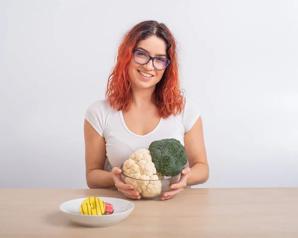 Kaukasiska kvinnor föredrar hälsosam mat. rödhårig flicka väljer mellan broccoli och munkar på vit bakgrund. — Stockfoto