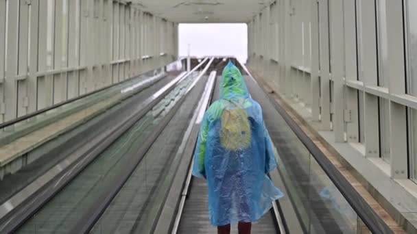 Uma mulher de gabardina sobe numa escada rolante. Menina em roupas protetoras da chuva. Visão traseira. — Vídeo de Stock