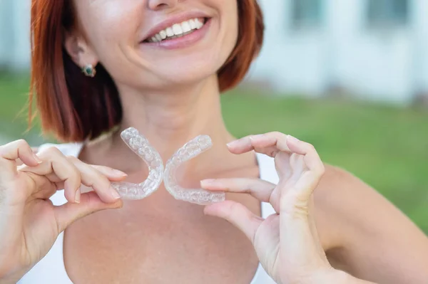 Mujer caucásica pelirroja sosteniendo protectores bucales transparentes para corrección de mordeduras al aire libre. Una chica con una hermosa sonrisa blanca como la nieve usa frenos de silicona — Foto de Stock