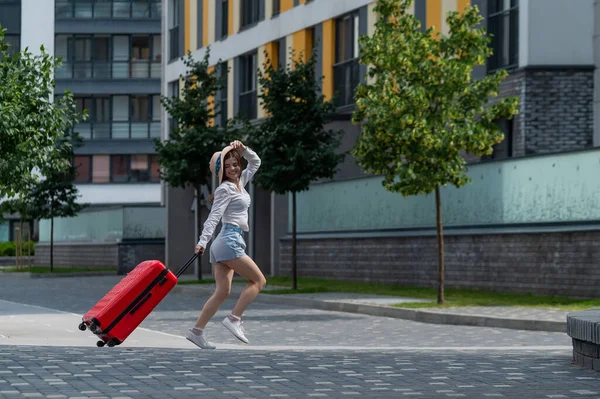 Glückliche kaukasische junge Frau mit Hut und kurzen Hosen hält einen großen roten Koffer in der Hand läuft auf einer Straße der Stadt — Stockfoto