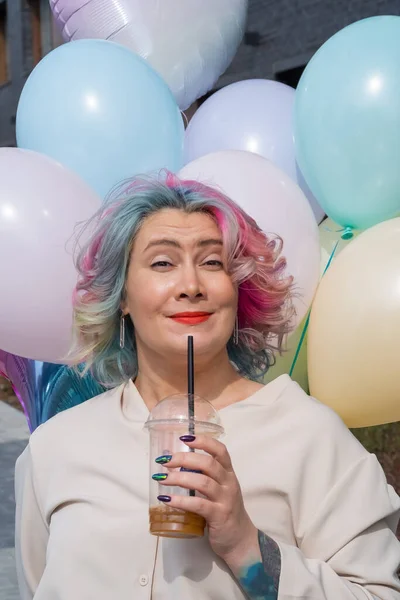 Žena v barevných vlasech chodí s náručí plnou balónků a pije osvěžující nápoj — Stock fotografie
