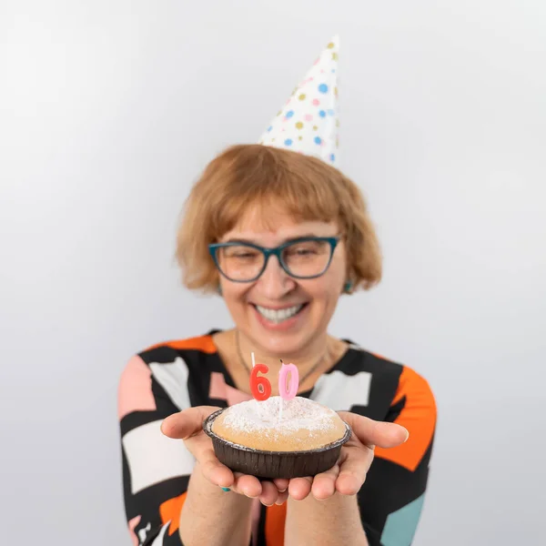 Ett porträtt av en leende äldre kvinna i en festlig mössa som håller en tårta med ljus i form av siffran 60. Årsdagen — Stockfoto