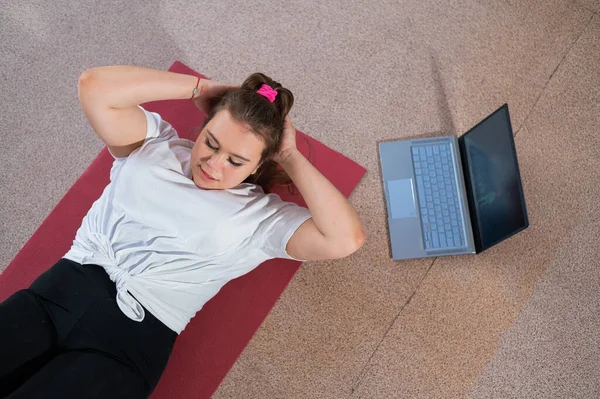 Μια παχουλή νεαρή γυναίκα παρακολουθεί ένα διαδικτυακό μάθημα γυμναστικής σε ένα λάπτοπ. Προπόνηση εξ αποστάσεως. — Φωτογραφία Αρχείου
