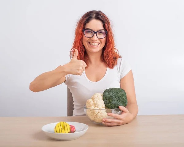 Kaukasiska kvinnor föredrar hälsosam mat. rödhårig flicka väljer mellan broccoli och munkar på vit bakgrund. — Stockfoto