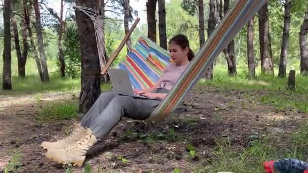 Mujer caucásica que trabaja en el ordenador portátil mientras está sentado en una hamaca en el bosque. Chica utiliza un ordenador inalámbrico en una caminata. — Vídeos de Stock