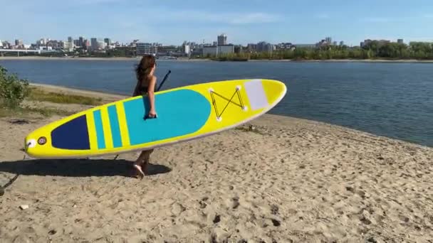 Mulher caucasiana caminha ao longo da praia e carrega uma prancha de jantar no rio na cidade. Desporto de verão. — Vídeo de Stock