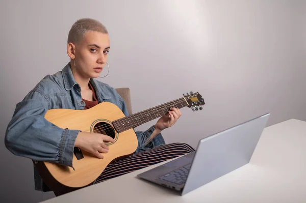 Jonge blanke vrouw met kort blond haar gitaar spelen en kijken naar training video op laptop. — Stockfoto