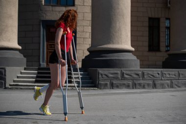 Şortlu genç kızıl saçlı kadın dışarıda koltuk değnekleriyle dolaşıyor..