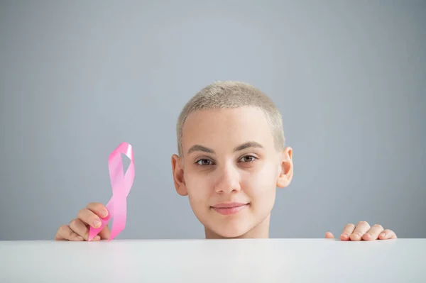 Young woman with short hair wearing a white t-shirt holding a pink ribbon as a symbol of breast cancer on a white background. — Stock Photo, Image