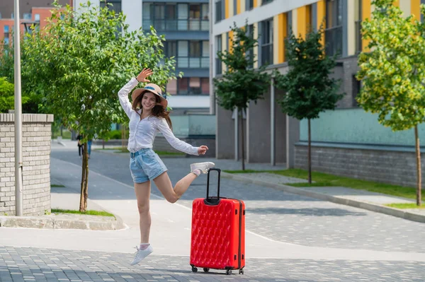 Gelukkig blanke jonge vrouw met een hoed en korte broek houdt een grote rode koffer vast loopt op een straat in de stad — Stockfoto