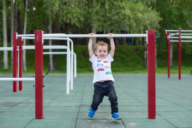 Küçük bir çocuk açık havada yatay bir çubukta durmayı öğreniyor. Çocuk dengesiz parmaklıklara asılmış..