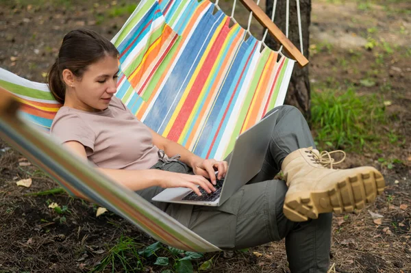 Mulher caucasiana trabalhando em laptop enquanto sentada em uma rede na floresta. Menina usa um computador sem fio em uma caminhada. — Fotografia de Stock