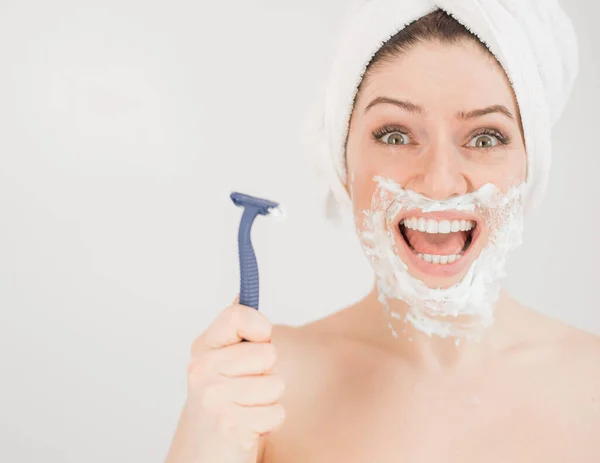 Cheerful caucasian woman with a towel on her head and shaving foam on her face holds a razor on a white background — Stock Photo, Image