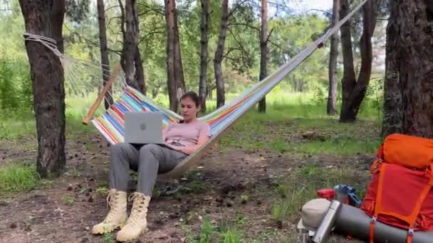 Vit kvinna som arbetar på laptop medan hon sitter i en hängmatta i skogen. Flicka använder en trådlös dator på en vandring. — Stockvideo
