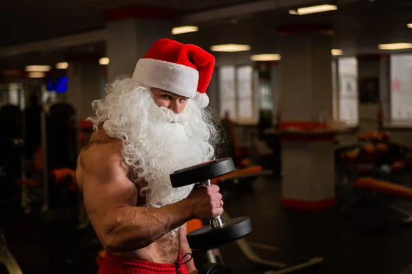Санта Клаус в спортзале. Мускулистый мужчина с обнаженным туловищем делает упражнения с гантелями. — стоковое фото