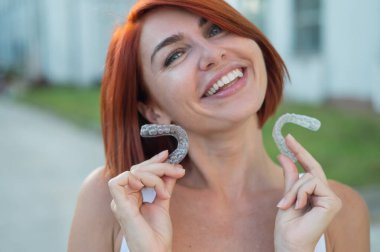 Kızıl saçlı beyaz kadın, ısırık düzeltmesi için şeffaf ağız koruyucuları tutuyor. Güzel kar beyazı gülüşlü bir kız silikon diş teli kullanır.