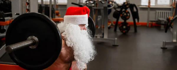 Santa Claus en el gimnasio. Hombre musculoso con un torso desnudo haciendo ejercicios con pesas. — Foto de Stock