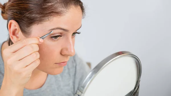 Кавказька жінка дивиться у дзеркало і виправляє брови за допомогою щипців. — стокове фото