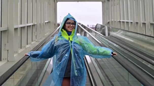 Mujer joven sonriente con chaqueta y impermeable en un travolator. Chica con ropa protectora de la lluvia — Vídeos de Stock