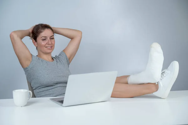 Femme caucasienne a soulevé sa jambe avec du plâtre au bureau et travaille sur ordinateur portable sur fond blanc — Photo