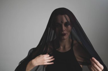 Karanlık bir stüdyoda siyah peçeli kızıl saçlı bir kadın. Burnunda küpe olan bir kız sinsice gülümsüyor. Cadı makyajı. Cadılar Bayramı kostümü.