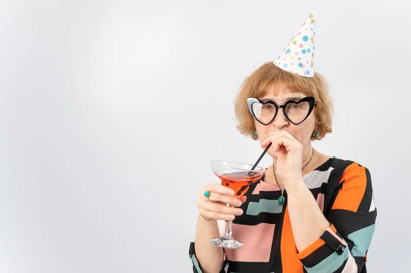 Porträtt av en leende äldre kvinna i en festlig mössa och glasögon med hjärtan dricka en cocktail på en vit bakgrund — Stockfoto