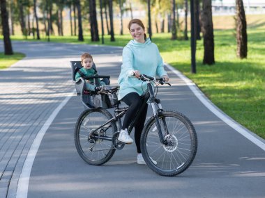 Küçük oğluyla bisiklete binen beyaz bir kadın arkasında çocuk koltuğunda oturuyor.