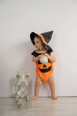 Beyaz arka planda kafatası tutan Cadılar Bayramı için balkabağı kostümü giymiş şirin beyaz çocuk.