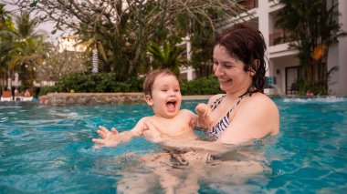 Beyaz bir kadın tatilde havuzda yüzerken oğlunu kucağına alıyor. Annesi küçük çocuğa yüzmeyi öğretiyor.