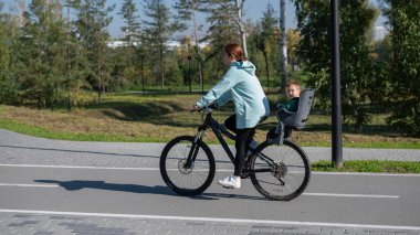 Beyaz kadın parkta bir yaşındaki oğluyla bisiklete biniyor.