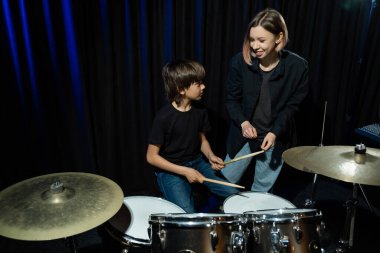 Genç beyaz bir kadın, bir çocuğa stüdyoda siyah arka planda davul çalmayı öğretiyor. Müzik okulu öğrencisi.