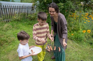 Beyaz kadın iki oğluyla barbekü yaparken ızgara sosis yapıyor.