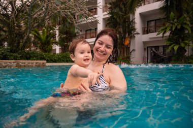 Beyaz bir kadın tatilde havuzda yüzerken oğlunu kucağına alıyor. Annesi küçük çocuğa yüzmeyi öğretiyor.