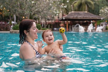 Beyaz bir kadın tatilde havuzda yüzerken oğlunu kucağına alıyor. Annesi küçük çocuğa yüzmeyi öğretiyor.