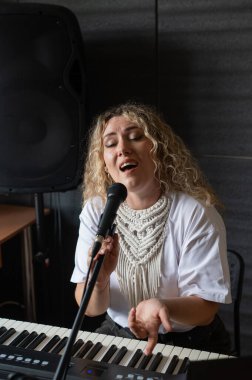 Beyaz kadın mikrofona şarkı söylüyor, sentezleyici çalıyor ve kendini dizüstü bilgisayarına kaydediyor.