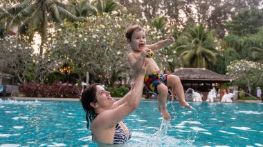 Beyaz bir kadın tatilde havuzda yüzerken oğlunu kucağına alıyor. Annesi küçük çocuğa yüzmeyi öğretiyor.
