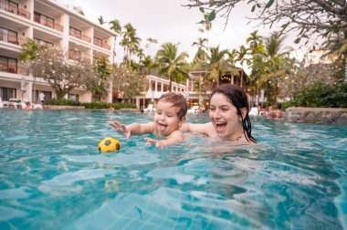 Beyaz bir kadın tatilde havuzda yüzerken oğlunu kucağına alıyor. Annesi küçük çocuğa yüzmeyi öğretiyor.