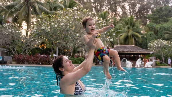 Beyaz bir kadın tatilde havuzda yüzerken oğlunu kucağına alıyor. Annesi küçük çocuğa yüzmeyi öğretiyor.